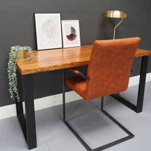 May include: A wooden desk with black metal legs and a brown leather chair. The desk has a natural wood finish and is decorated with two framed prints and a potted plant.