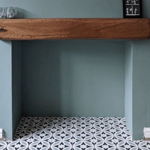 May include: A wooden mantel shelf with a light brown finish sits above a fireplace with a white and black patterned tile floor. The wall is painted a light blue-green color.