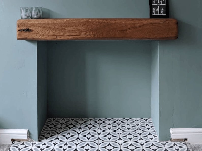 May include: A wooden mantel shelf with a light brown finish sits above a fireplace with a white and black patterned tile floor. The wall is painted a light blue-green color.
