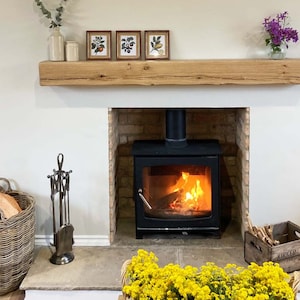 May include: A wood-burning fireplace with a black cast iron stove and a brick surround. The fireplace has a wooden mantel with three framed prints of botanical illustrations. A wicker basket filled with logs sits on the floor next to a set of fireplace tools.