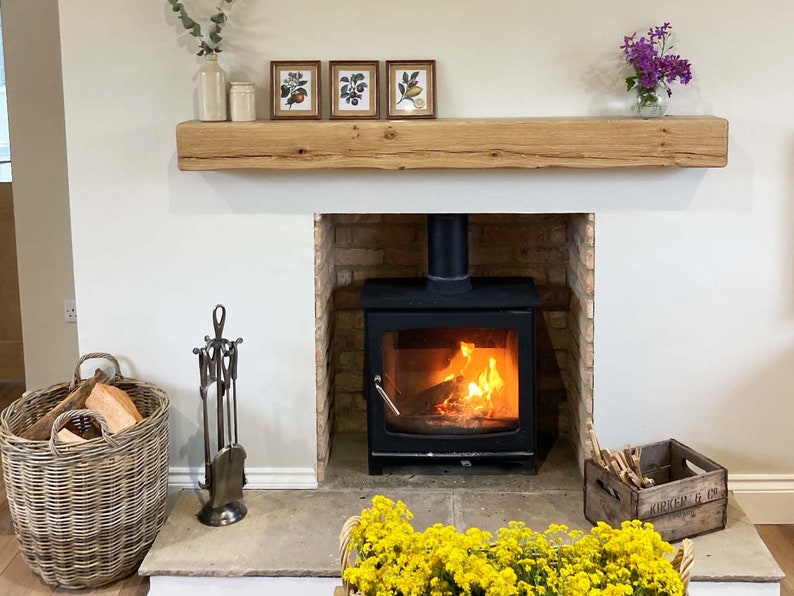 May include: A wood-burning fireplace with a black cast iron stove and a brick surround. The fireplace has a wooden mantel with three framed prints of botanical illustrations. A wicker basket filled with logs sits on the floor next to a set of fireplace tools.