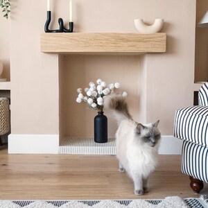 May include: A white cat with gray markings walks in front of a fireplace with a wooden mantel and a black vase with white flowers. The fireplace is in a room with light beige walls and a striped couch.