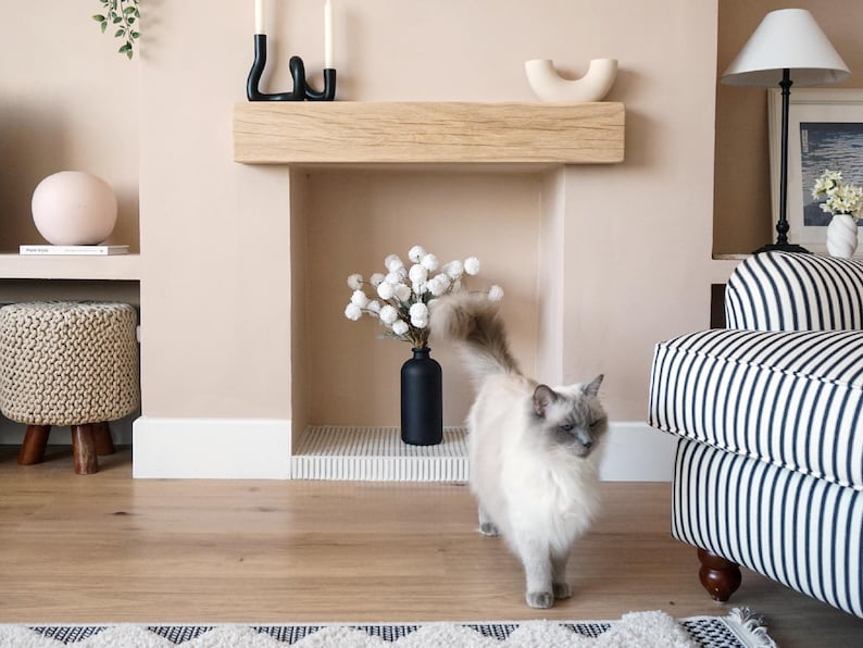 May include: A white cat with gray markings walks in front of a fireplace with a wooden mantel and a black vase with white flowers. The fireplace is in a room with light beige walls and a striped couch.