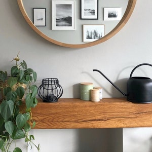 May include: A wooden shelf with a black watering can, three small jars, and a black metal candle holder. A potted plant with green leaves hangs over the shelf.