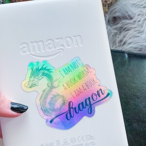 May include: A holographic sticker featuring a green dragon design and the text "I am not a book worm, I am a book dragon". The sticker is applied to a white Amazon Kindle.