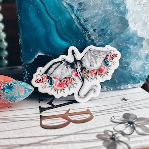 May include: A white sticker featuring a black and grey dragon silhouette surrounded by pink, red and blue flowers. The sticker is on a blue and white agate stone.