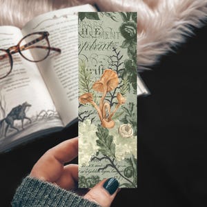 May include: A rectangular bookmark featuring a light green background with a vintage-style botanical illustration of orange mushrooms, dark green foliage, and black cursive script text layered over the design in a detailed, artistic composition.