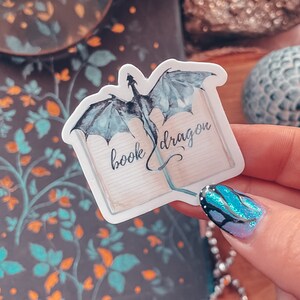 May include: A white sticker featuring a blue watercolour dragon emerging from an open book. The book is inscribed with the words "book dragon".