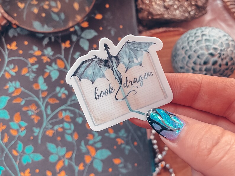 May include: A white sticker featuring a blue watercolour dragon emerging from an open book. The book is inscribed with the words "book dragon".