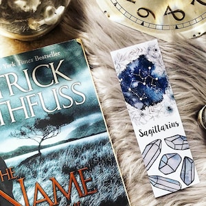 May include: A blue and white watercolor bookmark with the constellation Sagittarius and the text "Sagittarius" printed on it. The bookmark also features a floral design and several white crystals.