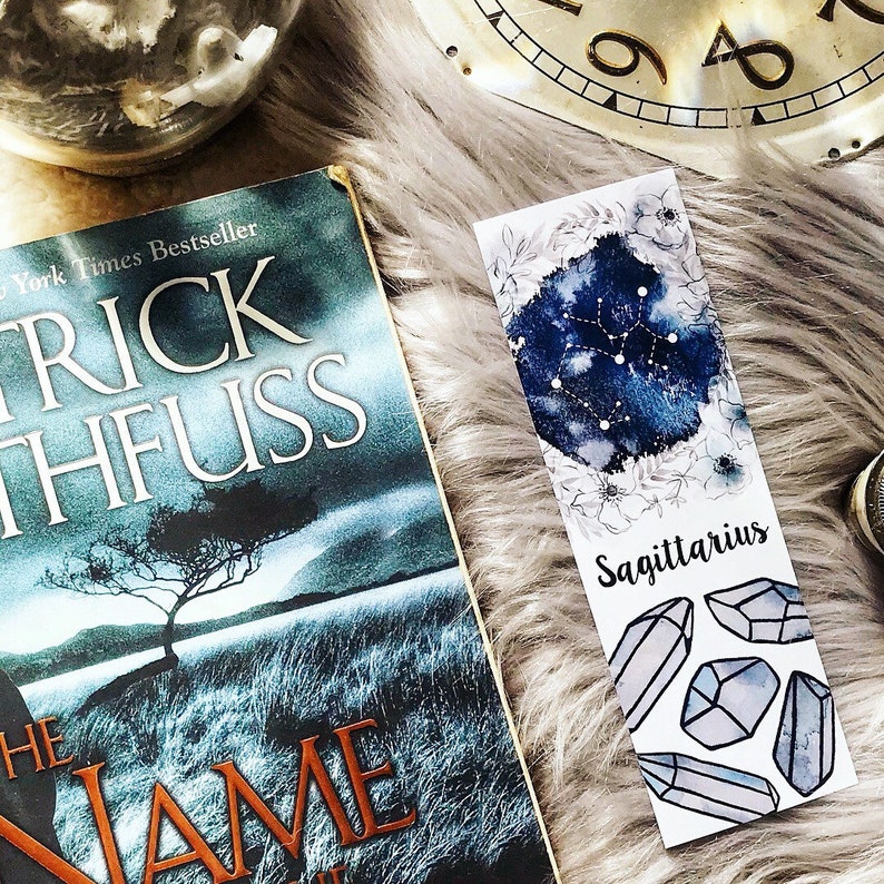 May include: A blue and white watercolor bookmark with the constellation Sagittarius and the text "Sagittarius" printed on it. The bookmark also features a floral design and several white crystals.