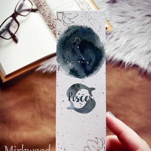 May include: A white bookmark with a watercolour design featuring the Pisces zodiac symbol. The top has a dark blue watercolour circle with the Pisces constellation in white. The bottom has the word "Pisces" and two fish. The bookmark is held by a person.