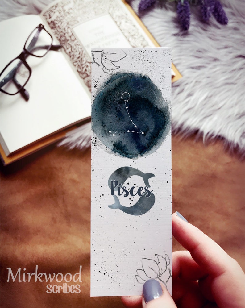 May include: A white bookmark with a watercolour design featuring the Pisces zodiac symbol. The top has a dark blue watercolour circle with the Pisces constellation in white. The bottom has the word "Pisces" and two fish. The bookmark is held by a person.