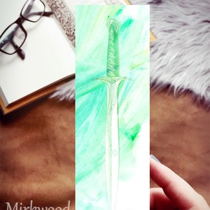 May include: A watercolor bookmark featuring a green sword with intricate details. The background is a soft green and yellow watercolor wash.