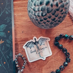 May include: A white sticker with a black and grey dragon flying out of an open book. The text on the sticker reads "book dragon".
