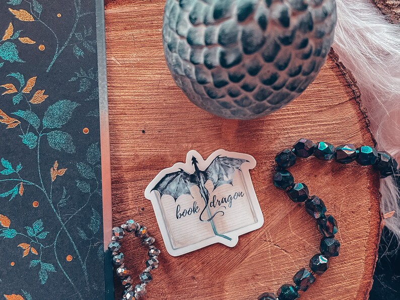 May include: A white sticker with a black and grey dragon flying out of an open book. The text on the sticker reads "book dragon".