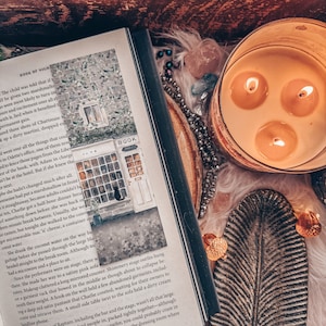 Peut inclure: Un livre ouvert avec une illustration aquarelle d'un b&acirc;timent avec une enseigne qui indique "BOOK". Le livre est ouvert &agrave; une page avec du texte sur un enfant qui re&ccedil;oit deux guimauves. Le livre est pos&eacute; sur une table avec une bougie allum&eacute;e et d'autres objets d&eacute;coratifs.