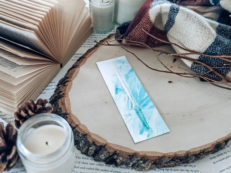 May include: A watercolor bookmark featuring a blue and silver sword with a white background.