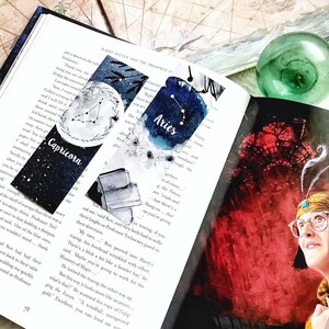 May include: Two watercolor bookmarks are displayed in an open book. One bookmark features the Capricorn constellation, and the other displays the Aries constellation. The bookmarks have a dark blue and white color scheme.