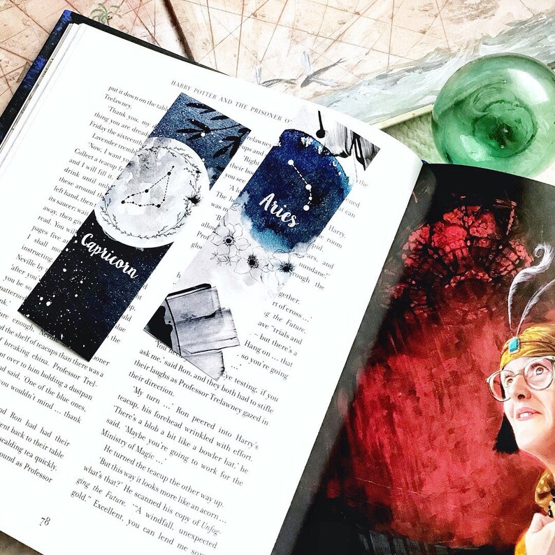May include: Two watercolor bookmarks are displayed in an open book. One bookmark features the Capricorn constellation, and the other displays the Aries constellation. The bookmarks have a dark blue and white color scheme.