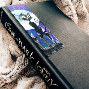 Peut inclure: Un livre reli&eacute; noir avec le titre "A Curse So Dark and Lonely" sur le dos. Le livre a un signet violet et bleu avec une illustration de chat noir.