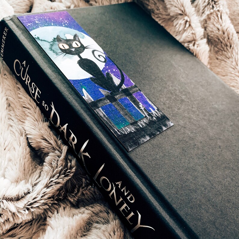 Peut inclure: Un livre reli&eacute; noir avec le titre "A Curse So Dark and Lonely" sur le dos. Le livre a un signet violet et bleu avec une illustration de chat noir.