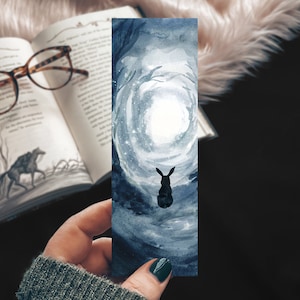 May include: A watercolor bookmark featuring a dark blue and gray forest scene with a white glowing circle in the distance. A black silhouette of a rabbit sits in the foreground.