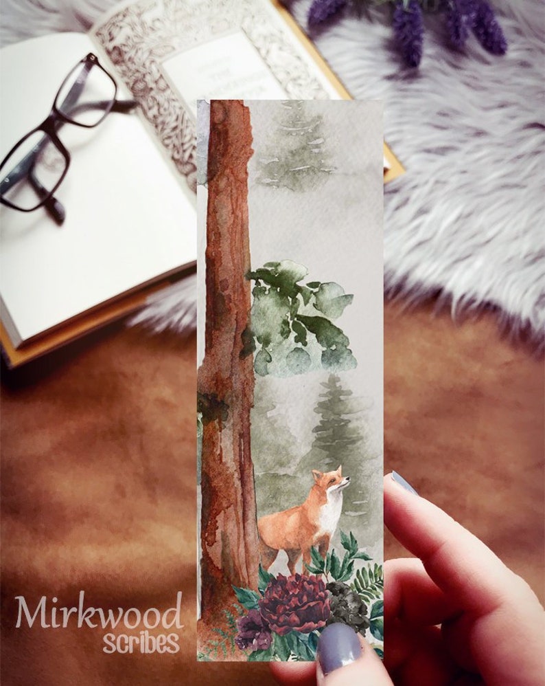 May include: A watercolor bookmark featuring a fox standing in a forest with a tree trunk and green foliage. The fox is orange and white with a bushy tail. The background is a soft gray with hints of green and brown. The bookmark has the text "Mirkwood Scribes" at the bottom.