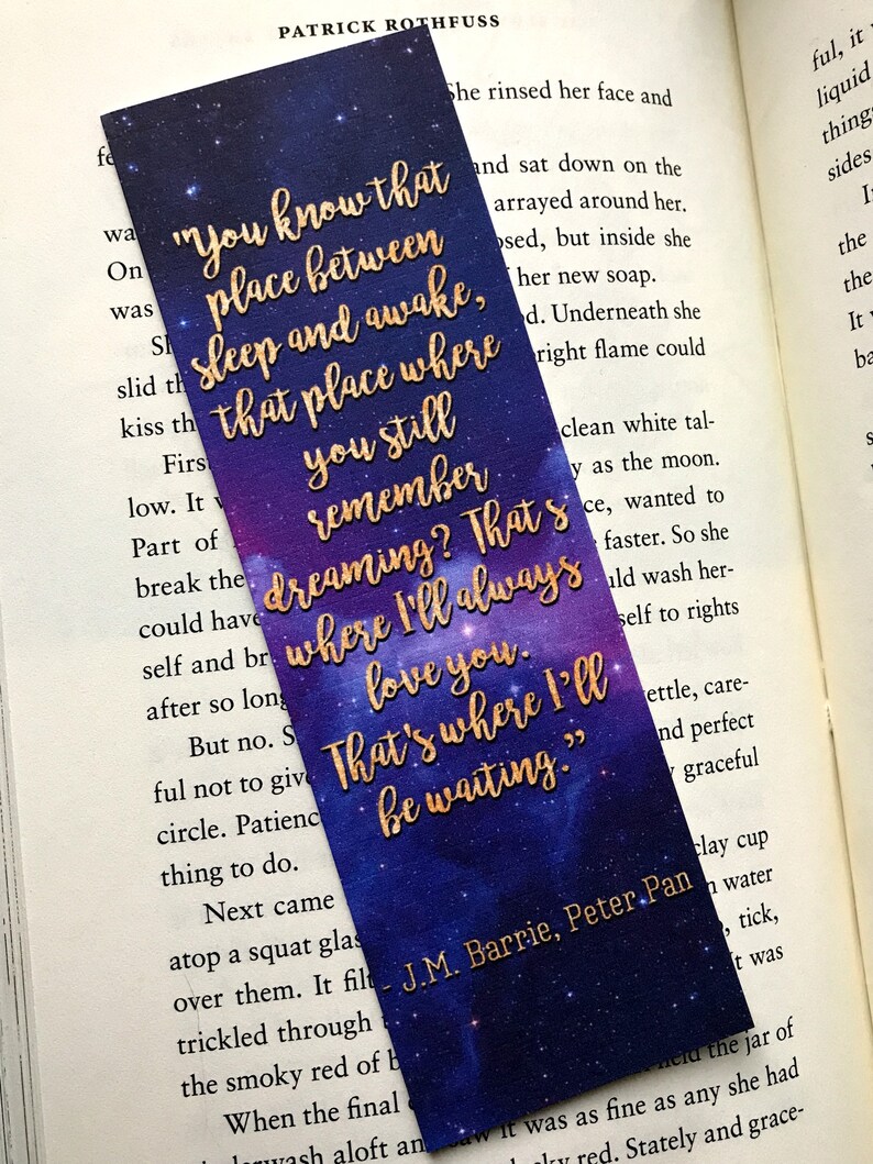 K&ouml;nnte beinhalten: Ein violettes und blaues Lesezeichen mit goldenen Glitzersternen und Text. Der Text lautet: "Du kennst diesen Ort zwischen Schlaf und Wachsein, diesen Ort, an dem du dich noch an das Tr&auml;umen erinnerst? Dort werde ich dich immer lieben. Dort werde ich warten." Das Lesezeichen enth&auml;lt auch den Text "J.M. Barrie, Peter Pan."