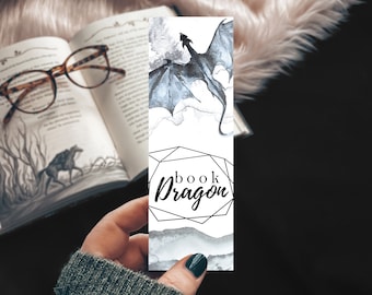 May include: A white bookmark featuring a black and grey watercolour dragon design. The bookmark has a geometric frame with the text "book dragon" inside.
