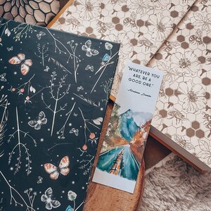 May include: A decorative bookmark with a watercolor landscape and the quote "WHATEVER YOU ARE, BE A GOOD ONE." by Abraham Lincoln. The bookmark rests on a wooden surface next to a book with a dark cover featuring butterflies and floral designs.