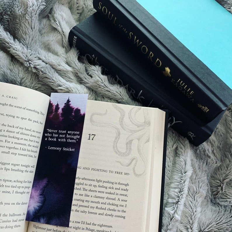 May include: A close-up of a bookmark with a watercolor illustration of trees in a forest. The bookmark has a quote from Lemony Snicket: "Never trust anyone who has not brought a book with them." The bookmark is placed on top of an open book with a drawing of an octopus.
