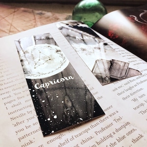 May include: Black and white bookmark with a silver constellation design and the word "Capricorn" in white lettering. The bookmark has a watercolor effect and a glittery finish.