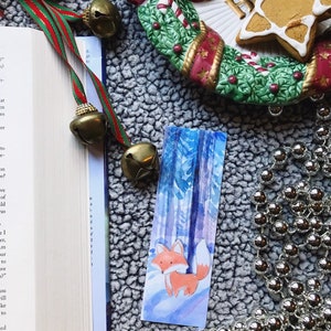 May include: A watercolor bookmark featuring a fox standing in a snowy forest. The fox is orange with a white tail and chest. The forest is painted in shades of blue and purple.