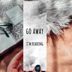 May include: A white bookmark with a gray watercolor background. The bookmark has black text that reads "GO AWAY I'M READING".