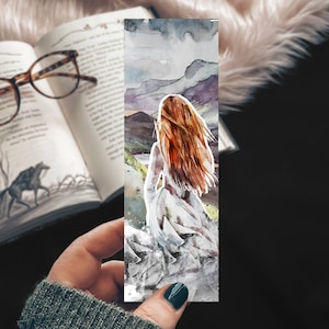 May include: Watercolor bookmark featuring a woman with long red hair standing on a cliff overlooking a landscape. The background is a watercolor wash of blue, gray, and purple.