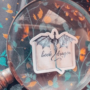 May include: A sticker featuring a black and white illustration of a dragon emerging from a book with the text "book dragon".