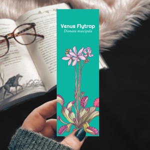 May include: A teal bookmark featuring a Venus Flytrap illustration with the text "Venus Flytrap Dionaea muscipula." The bookmark has a detailed botanical drawing of the plant with pink and green hues. The bookmark is held near an open book with glasses.