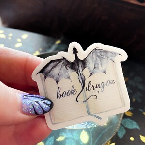 May include: A white sticker featuring a black and grey watercolour illustration of a dragon flying out of an open book. The text "book dragon" is written in cursive on the book.