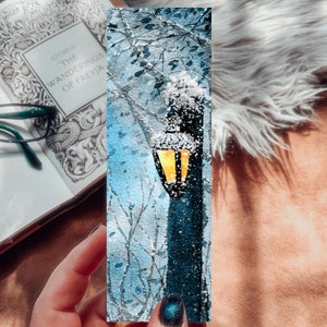 May include: A wintery scene with a street lamp covered in snow. The lamp is lit and casting a warm glow. The background is a blue and white watercolor painting of trees and snow.