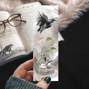 May include: A watercolour bookmark featuring a black crow flying over a branch with green leaves and orange berries. The background is a grey wash.