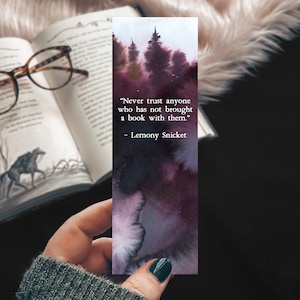 May include: A purple and black watercolor bookmark with a quote from Lemony Snicket: "Never trust anyone who has not brought a book with them."