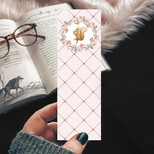 May include: A decorative bookmark featuring a watercolor illustration of a squirrel inside a floral wreath. The bookmark has a light pink background with a red diamond pattern. The bookmark is being held near an open book with glasses.