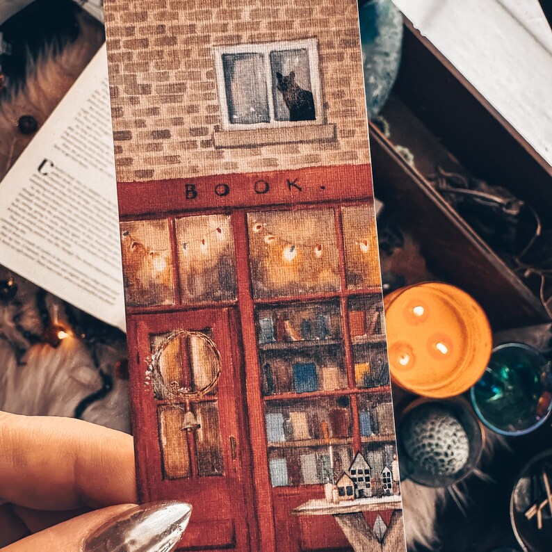 May include: A watercolor illustration of a storefront with a red door and a sign that reads "BOOK". The storefront has a window with a black cat looking out. The illustration is painted on a textured paper background.
