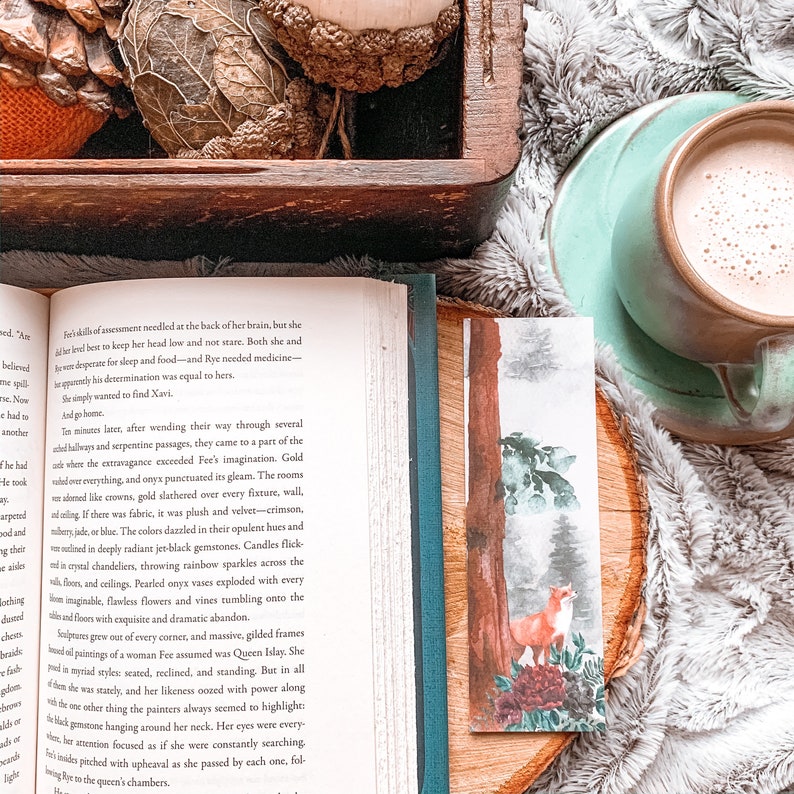 May include: A watercolor bookmark featuring a fox standing in a forest with a tall tree in the background. The fox is orange and white with a bushy tail. The forest is painted in shades of green and blue.