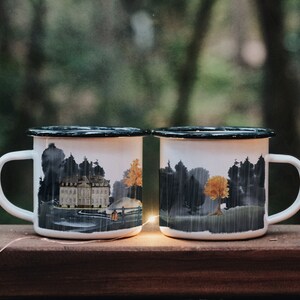 May include: Two white enamel mugs with black rims. The mug on the left has a watercolor illustration of a castle in a forest. The mug on the right has a watercolor illustration of a forest with a single yellow tree.