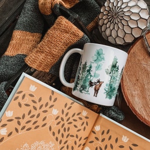 May include: A white ceramic mug with a watercolor design of a deer in a forest. The mug is sitting on a book with a patterned cover and a brown and green knitted scarf.