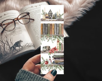 May include: A decorative bookmark featuring watercolor illustrations of cozy winter scenes. The bookmark showcases images of a miniature village, a shelf of books, candles, and festive holiday decorations. The bookmark is held in a hand, with an open book and glasses in the background.