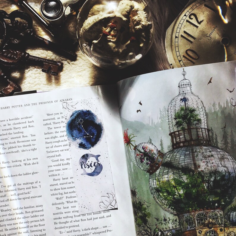May include: A detailed image featuring an open book with illustrations from "Harry Potter and the Prisoner of Azkaban", vintage keys, a pocket watch, and a glass terrarium. The book displays watercolor art and the word "Pisces".