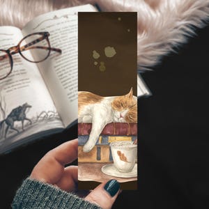 May include: Watercolor bookmark featuring a sleeping orange and white cat resting on a stack of books. A cup of coffee with a feather design sits below. The bookmark is held in a hand, with a book and glasses in the background.